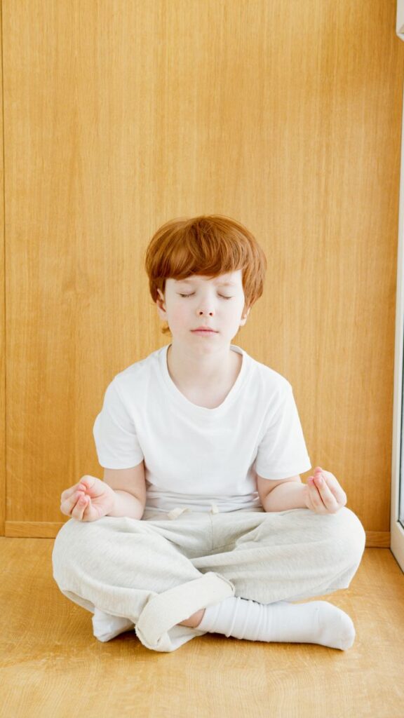 Young redheaded boy meditating indoors on a wooden floor, eyes closed in peaceful concentration.